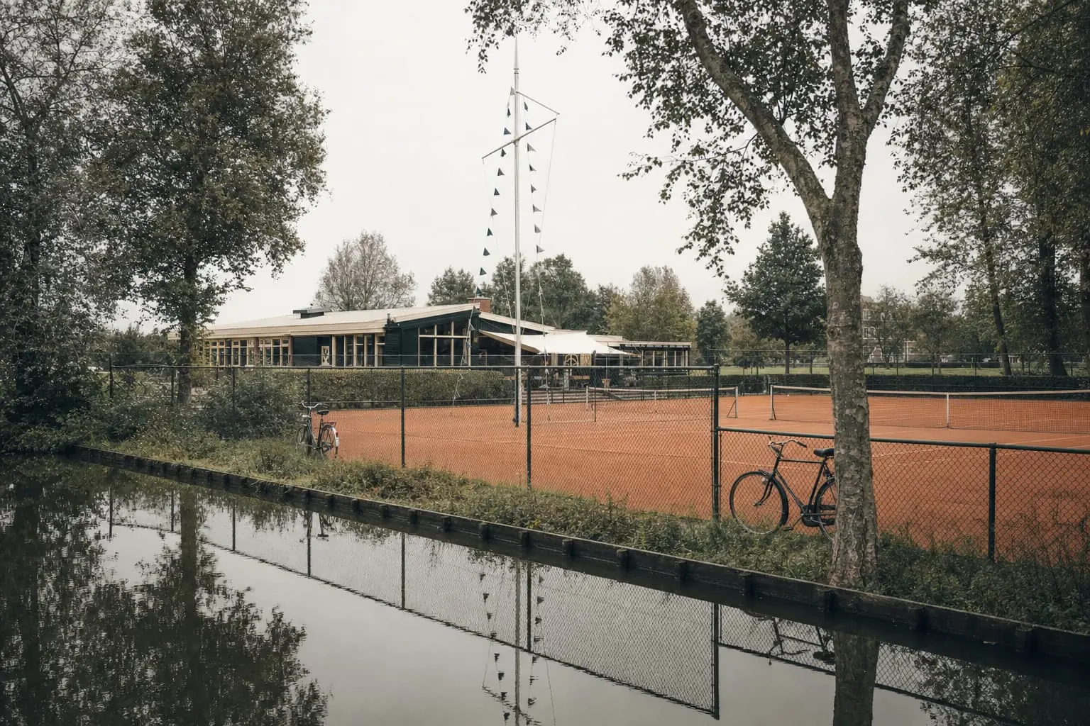 Gravel clay courts beside an Amsterdam canal, bicycles leaning on the court fence in flat polder light.