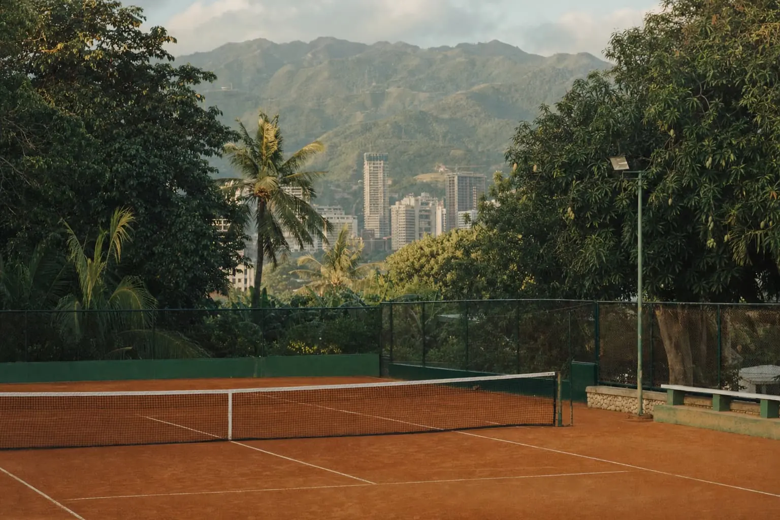 A clay court in Cebu framed by mango trees and palms, high-rise buildings and the green central mountains in the distance.