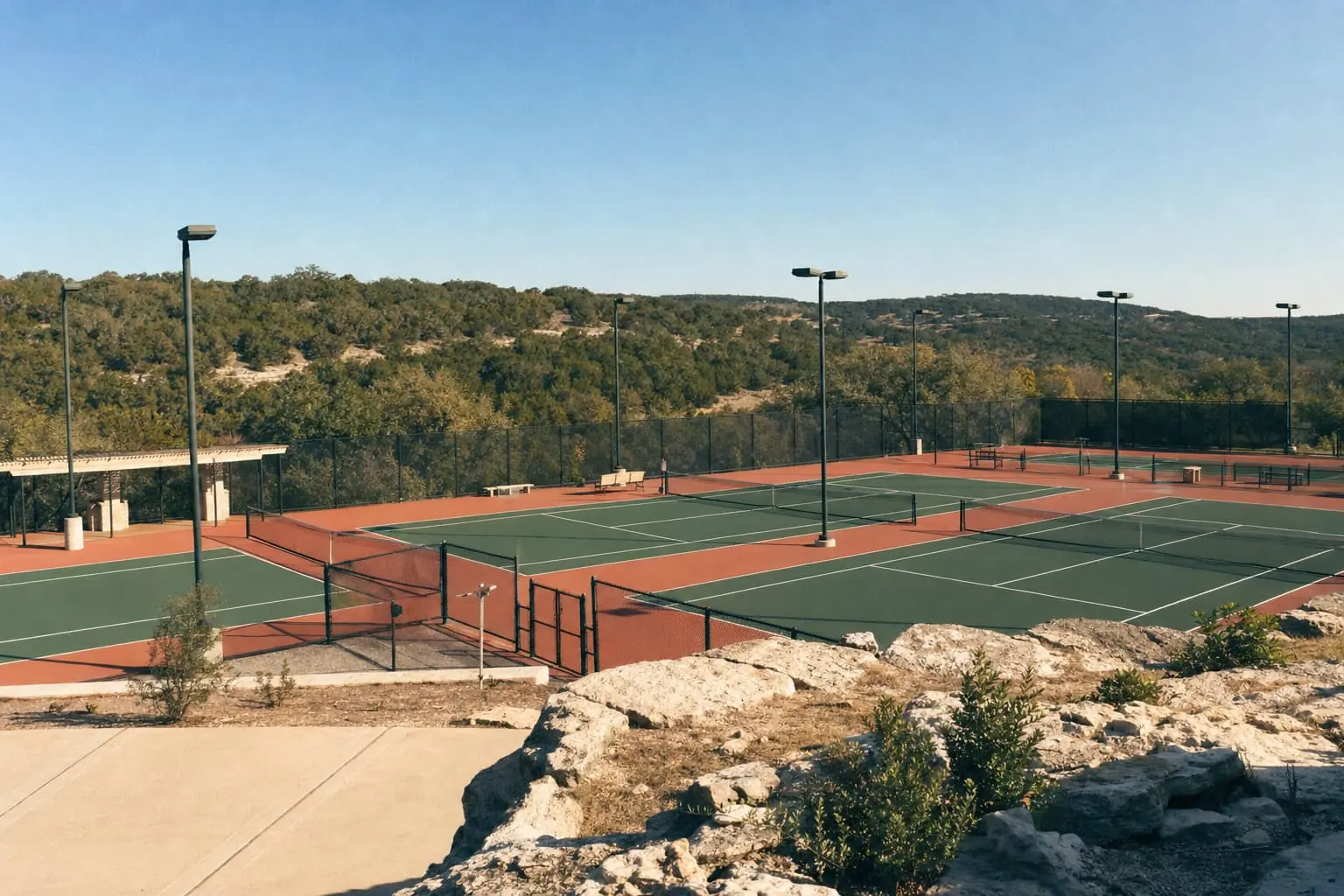 Floodlit public hard courts in Cedar Park on a Hill Country rise, juniper-covered hills and bare limestone rock under a clear blue sky.