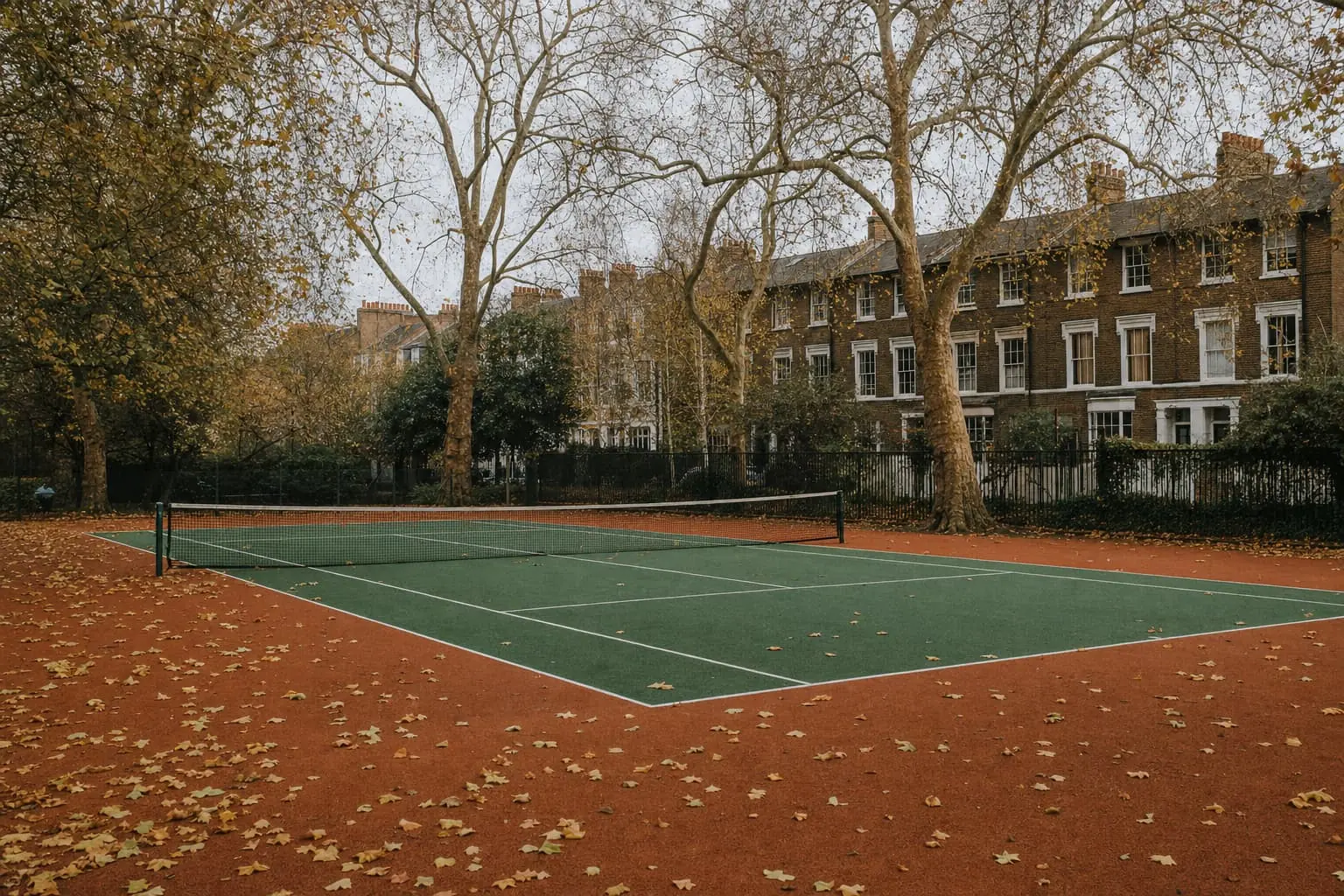 Autumn park hard courts in London, plane leaves on the lines, brick terraced houses beyond the railings.