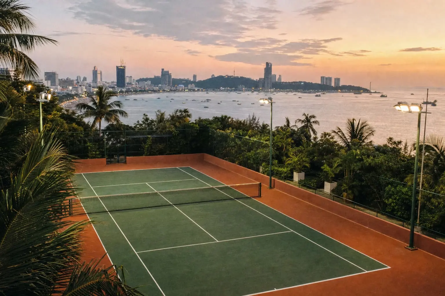 A floodlit hard court above Pattaya Bay at dusk, palms in the foreground and the city's towers along the curve of the bay.