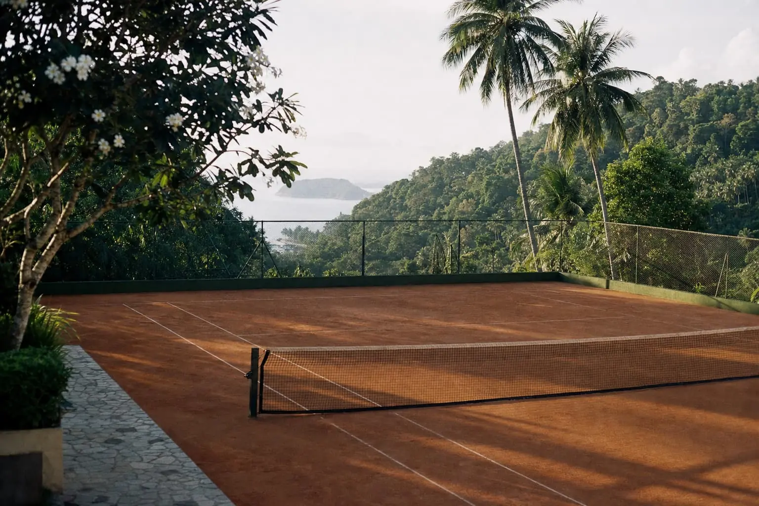 A hillside clay court on Phuket under coconut palms and a flowering frangipani, the Andaman Sea and an offshore island beyond.