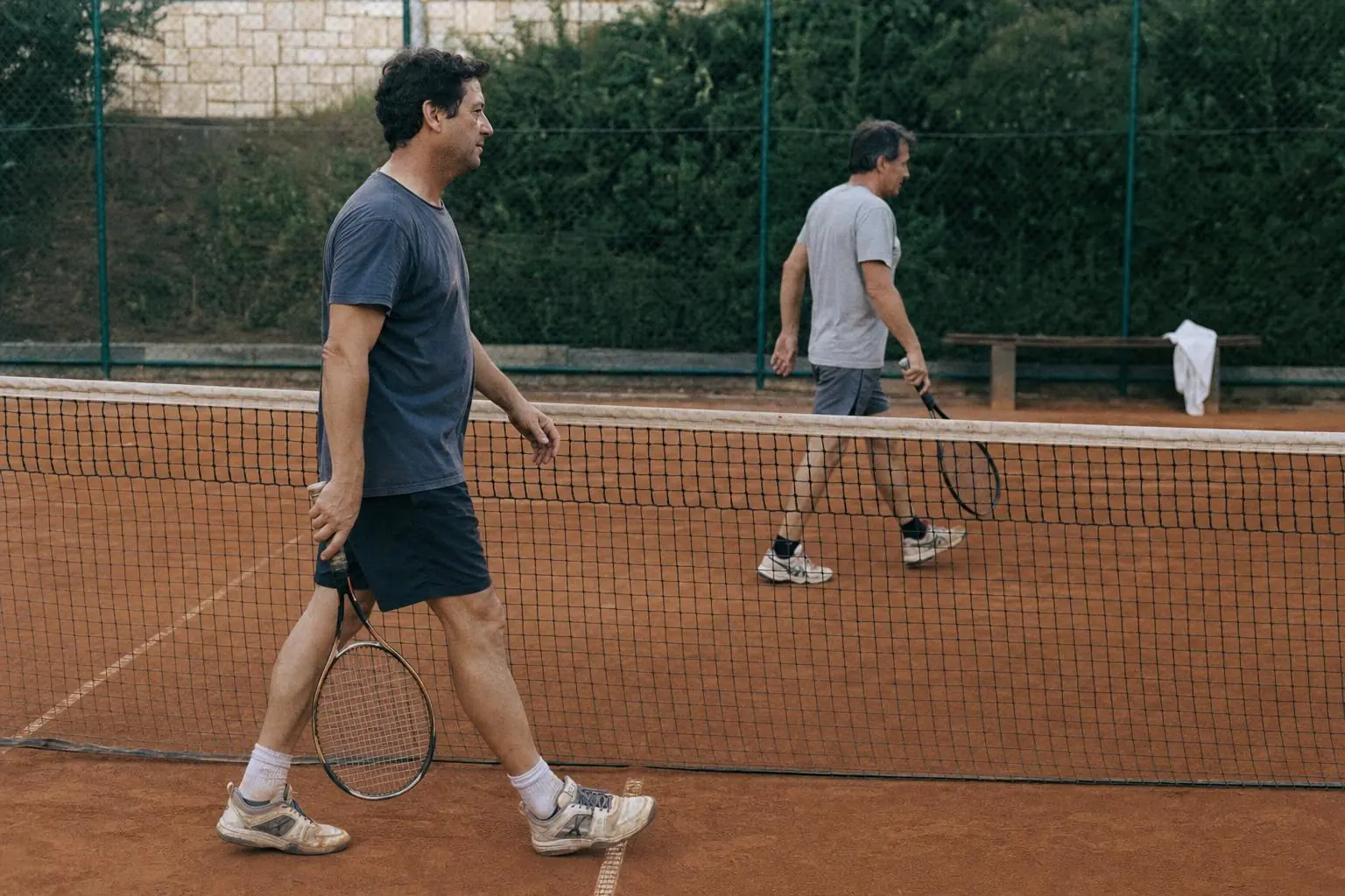 Two recreational tennis players walk beside the net on a worn clay court, carrying rackets, with a towel draped over a bench beyond the court.