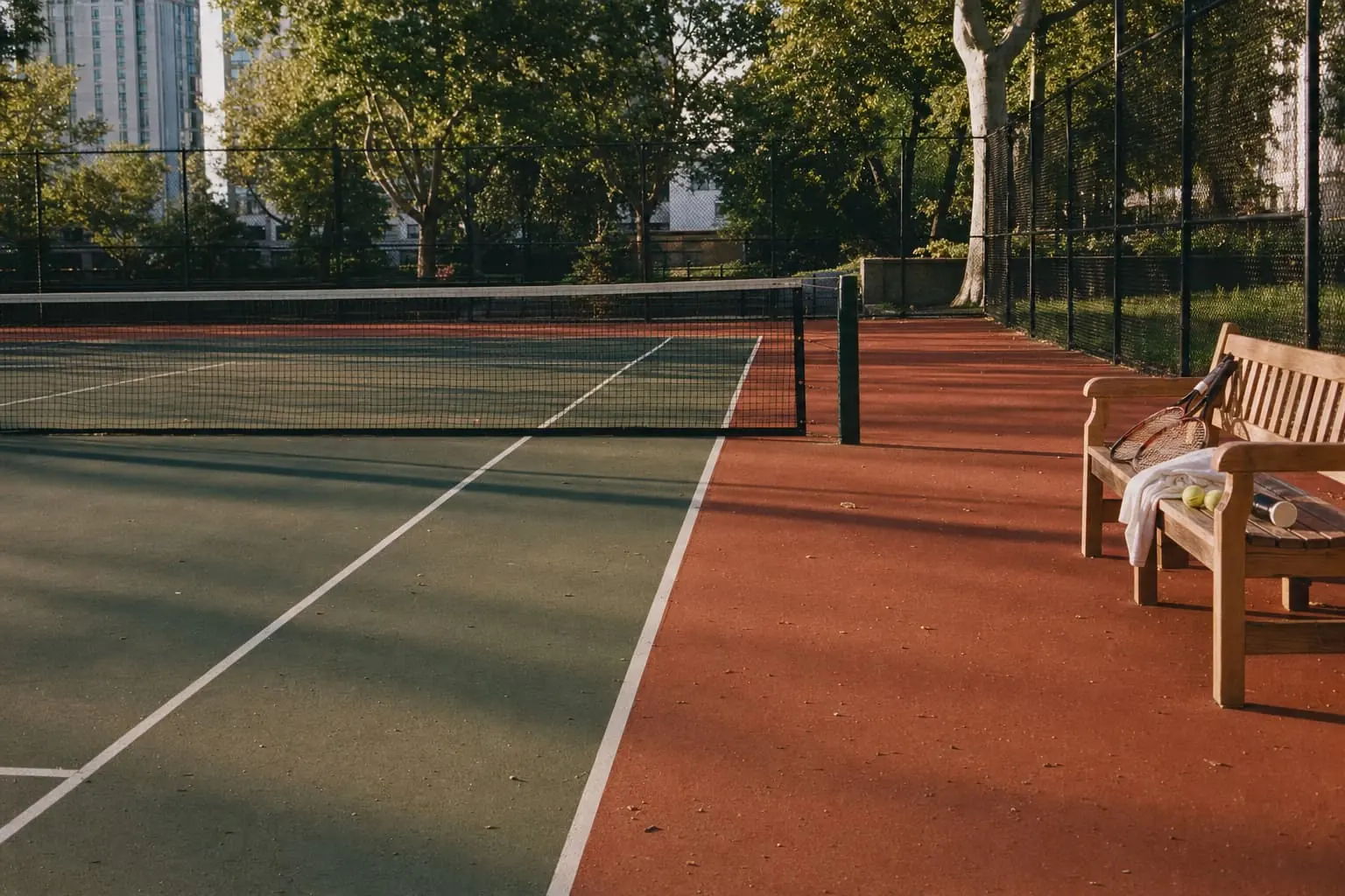 An empty green hard court with a terracotta apron in late-afternoon sun, the shadow of the net across it, and two rackets, a towel and balls on a wooden bench by the net post.