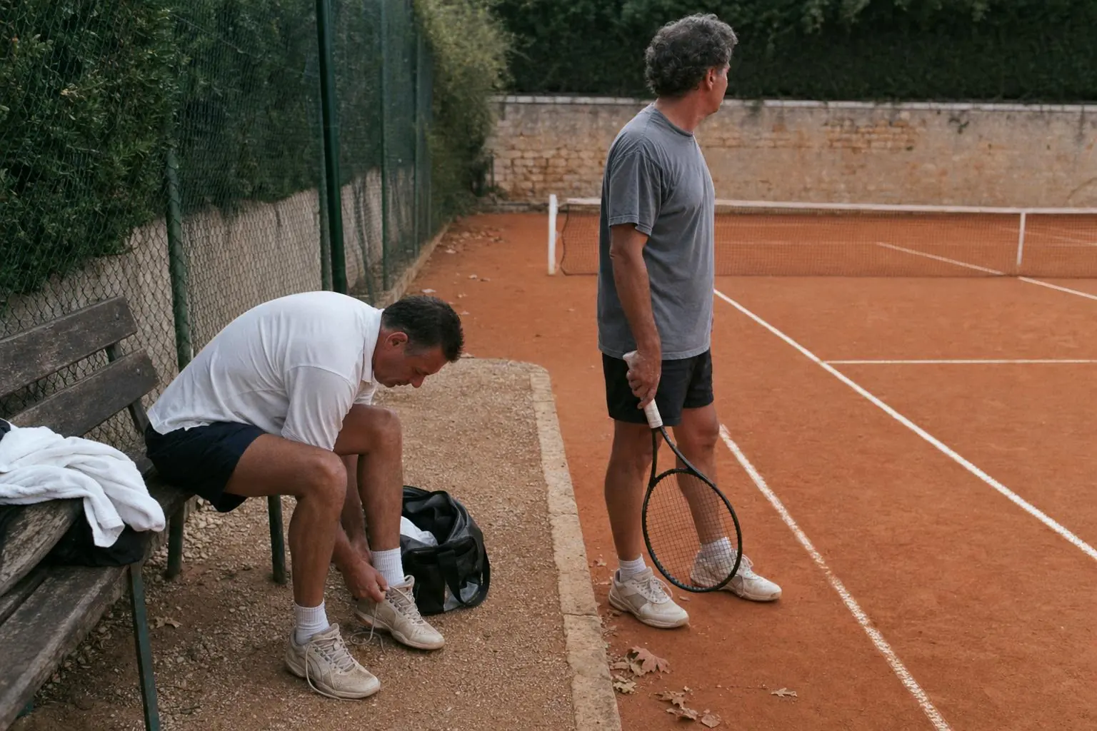 Two club players beside a clay tennis court, one bending over his shoes on a worn bench and the other holding a racket, with a towel and open bag nearby.