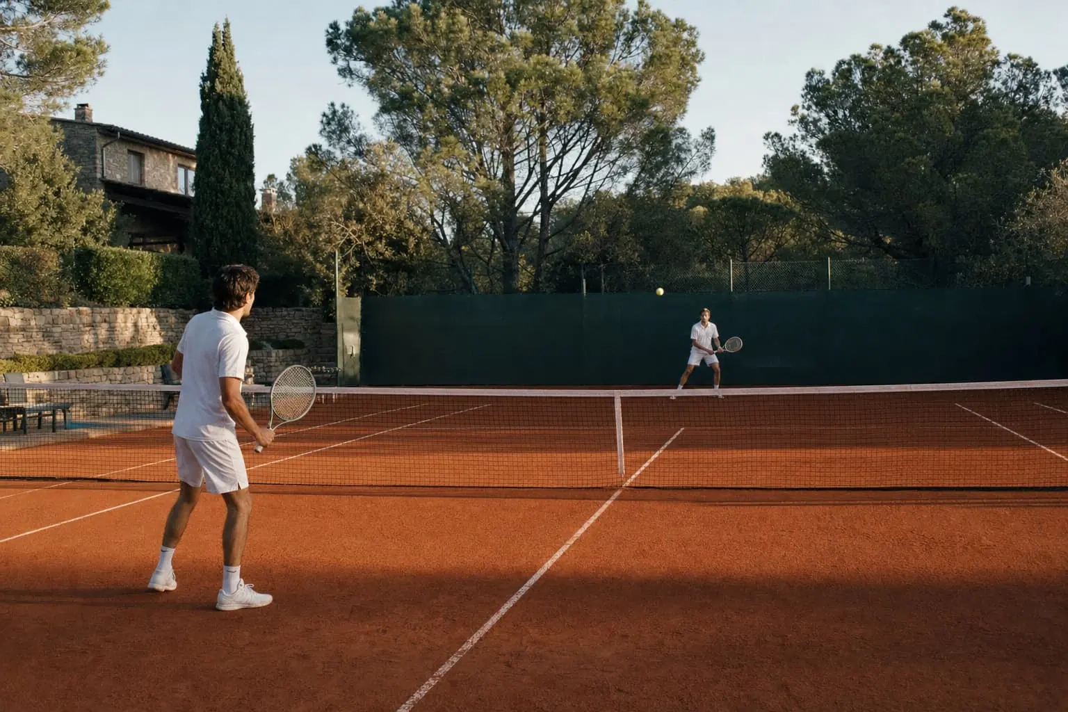 Two players warming up with a soft rally from the service lines of a clay court, pines and a stone house behind the fence.
